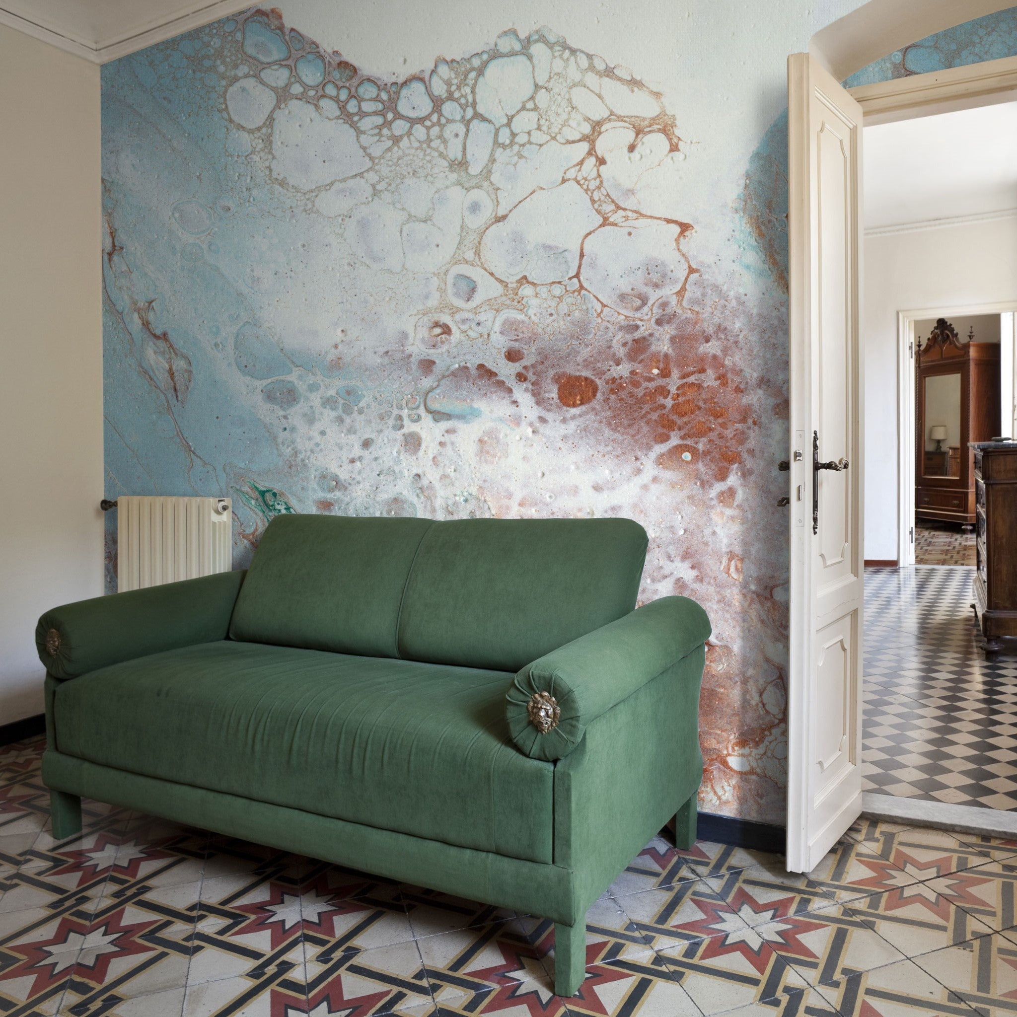 "Montage Wallpaper by Wall Blush accenting living room wall behind a green sofa, patterned tile flooring in view."