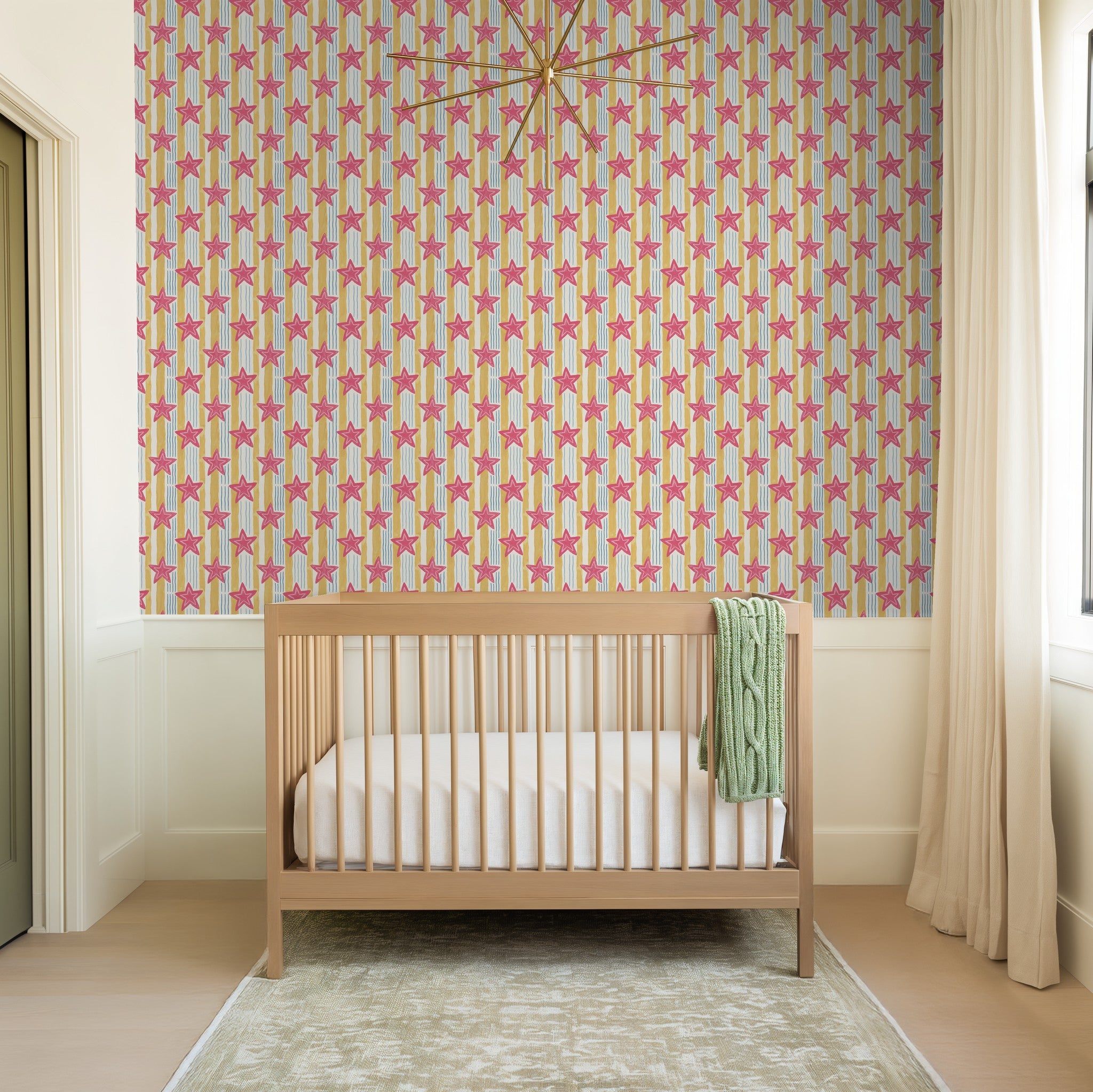 Starry Shore wallpaper with pink stars on yellow and blue stripes in a nursery with crib.